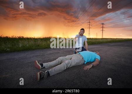 Young man calling emergency services for unconscious middle aged man. Dramatic resuscitation on rural road under storm clouds. Themes rescue, help and Stock Photo