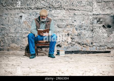 An older man reads a book, sits on an old chair, reads a book Stock Photo