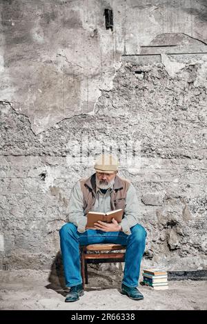 An older man reads a book, sits on an old chair, reads a book Stock Photo