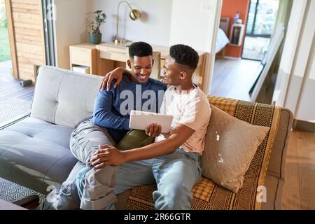 Happy young gay male couple hugging and using digital tablet on sofa Stock Photo