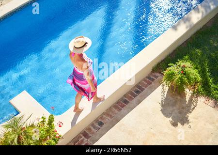 Woman in straw hat walking along edge of sunny swimming pool Stock Photo