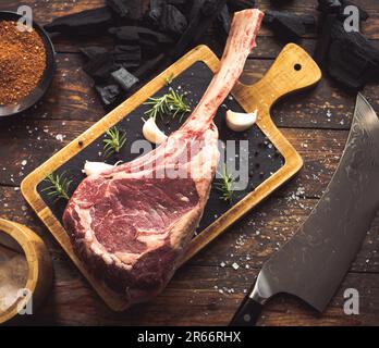 Dry aged raw tomahawk black angus prime beef chop with salt, pepper, rosemary, garlic, charcoal and a knife Stock Photo