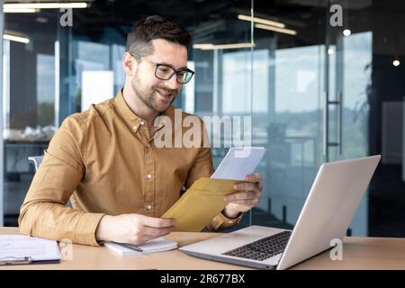 A young man businessman sits smiling in the office at the table and holds in his hands and reads documents, a contract, a successful deal that he received by mail. Stock Photo