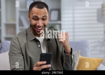 Close-up photo. Young hispanic man rejoices at the received message, reads good news, makes a victory gesture with his hand. Stock Photo