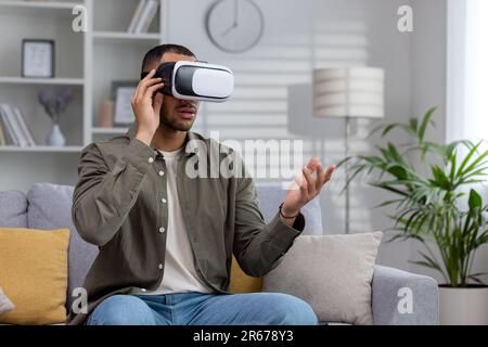 Young hispanic man wearing virtual glasses sits on the couch at home and watches online video through a mask. Gesturing with hands. Stock Photo