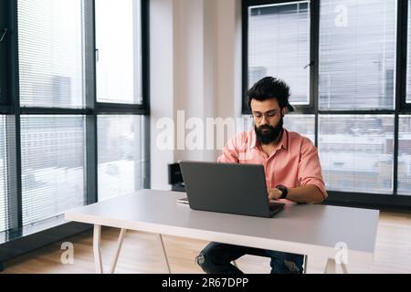 Concentrated Indian programmer male in eyeglasses working on laptop sitting at desk in light coworking office room by window. Stock Photo