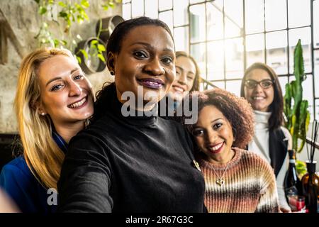 Multiethnic female friends taking selfie while smiling on camera in the modish office - Happy multicultural women having fun together - Bright filter Stock Photo