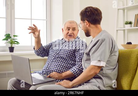 Friendly young man nurse helping senior person in using laptop sitting at home. Stock Photo