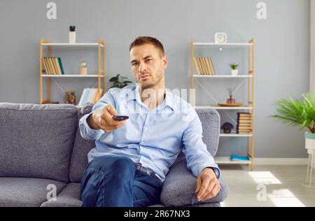Serious man sitting on sofa, watching TV, holding remote control and switching channels Stock Photo