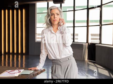 Mature Businesswoman Talking On Cell Phone Standing At Workplace Indoor Stock Photo