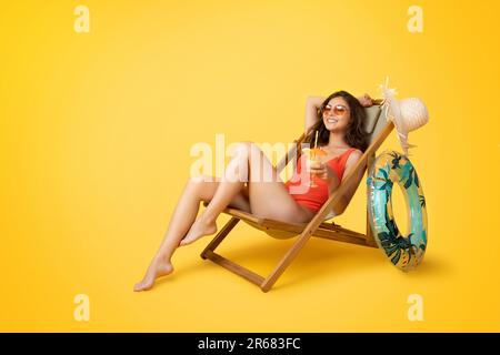Happy millennial mixed race woman in swimsuit sits in deck chair with tropical cocktail, enjoys holiday Stock Photo