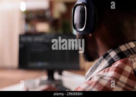 Closeup on freelance coder with wireless headphones listening to music while developing software on personal computer at home. African american programmer writing code while working remote on pc. Stock Photo