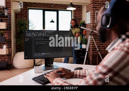 African american man with wireless headphones writing code on personal computer while girlfriend is using laptop standing in living room. Programmer developing software debugging algorithm at home. Stock Photo