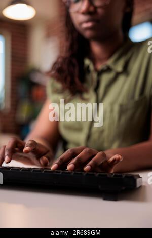 Closeup of african american woman programmer writing code on computer keyboard while working remote from home living room. Selective focus on hands typing and coding software on pc. Stock Photo