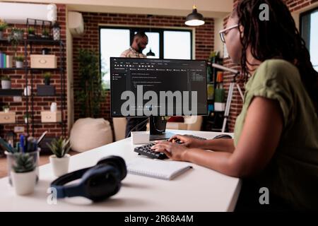 Woman coder developing software for freelance project working remote from home while boyfriend is relaxing. African american programmer writing source code looking for errors while debugging. Stock Photo