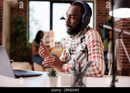 Freelancer with wireless headphones talking and explaining while doing hand gesture in video call conference on laptop. Coder attending internet meeting gesturing in front of portable computer. Stock Photo