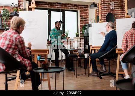 Friendly African American man teacher guiding group of different aged people through basics of drawing at art class. Young guy instructor explaining sketching techniques to students during workshop Stock Photo