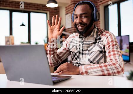 Programmer with wireless headphones in video conference waving hello at client while sitting at desk in living room. Smiling coder greeting friend in internet call about software development. Stock Photo