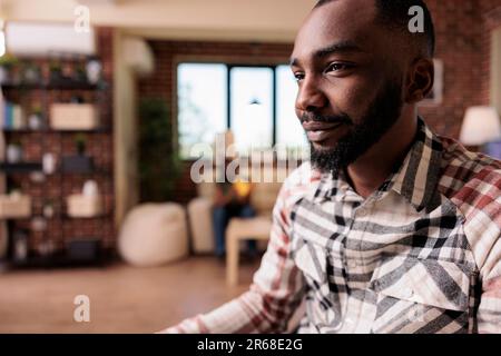 Closeup of african american freelancer reading content on computer screen while girlfriend is reading a book on the couch. Programmer working remote sitting at desk while roommate is relaxing on sofa. Stock Photo
