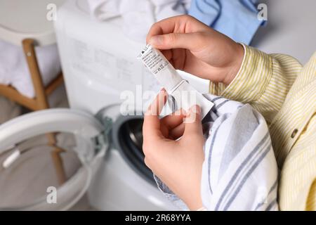 Woman reading clothing label with care symbols and material content on shirt near washing machine, closeup Stock Photo