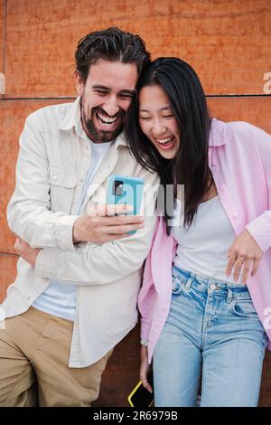 Vertical portrait of young adult multiracial couple having fun and laughing using a smartphone outside. Happy friends smiling and watching funny videos on a cellphone app. People using a mobile phone. High quality photo Stock Photo