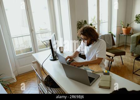 Focused male programmer sitting at desk and working at laptop and computer in home office Stock Photo