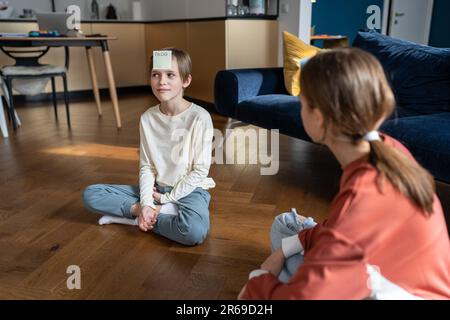 Smiling teen sister and child brother playing who am i game sitting on floor at home in living room. Stock Photo