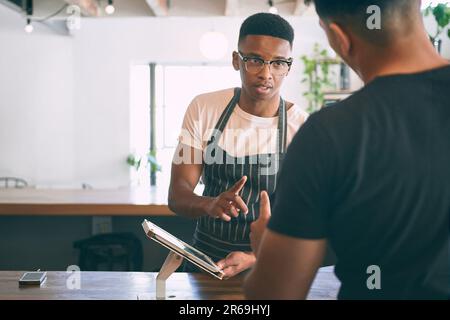How many sugars would you like with that. a young man using a digital tablet while buying coffee in a cafe. Stock Photo
