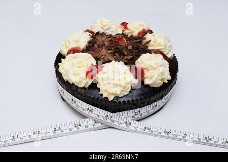 A measuring tape wrapped around a chocolate mud cake with whipped cream and jam to illustrate how too much junk food can cause weight gain, white background Stock Photo