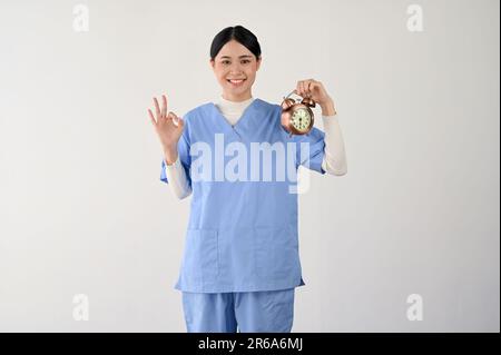 An attractive and smiling Asian female doctor in scrubs shows the Okay hand sign and holds a vintage alarm clock while standing against an isolated wh Stock Photo