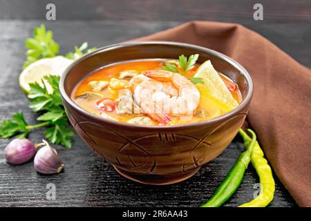 Thai soup Tom Yam from shrimp, chicken meat with mushrooms, tomatoes and lemon in a bowl, brown towel on a wooden board background Stock Photo