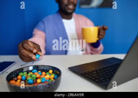 Closeup of black man enjoying candy snack and coffee while using laptop in vibrant office, copy space Stock Photo