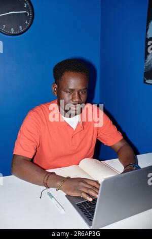 Vibrant vertical portrait of focused black man using laptop at workplace shot with flash Stock Photo