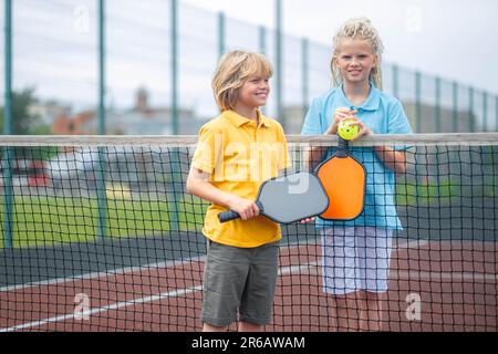 Portrait smiling pickleball game players boy and girl with yellow ball and paddles, outdoor sport leisure kids activity. Stock Photo