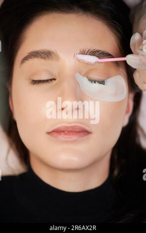 Woman with eye patch under lower eyelid keeping eyes closed while beautician applying cleansing mousse on eyebrows with lash brush. Female person having eyelash extension procedure in beauty salon. Stock Photo