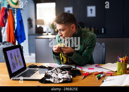 Biracial transgender fashion designer sitting at desk, using laptop and holding fabric Stock Photo