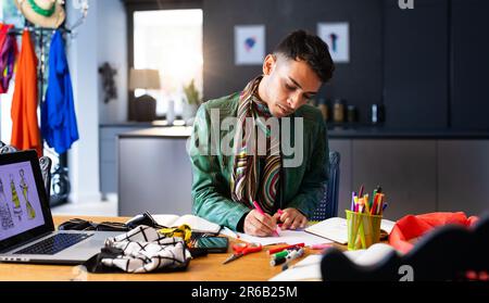 Biracial transgender fashion designer sitting at desk, using laptop and drawing sketches Stock Photo