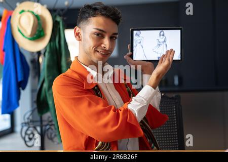 Happy biracial transgender fashion designer sitting at desk, holding tablet and having video call Stock Photo