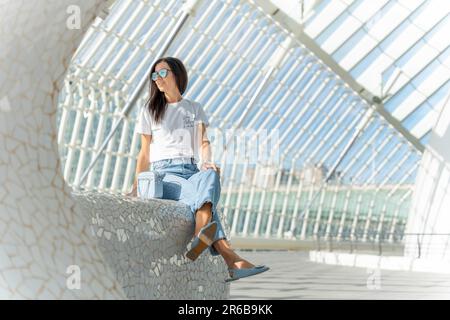 A young woman wearing eyeglasses is seated atop a grey cement bench Stock Photo
