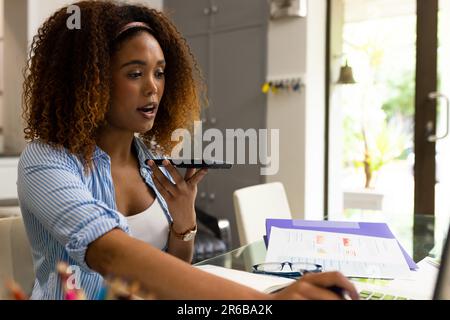 Busy biracial woman working at home, using laptop and talking on smartphone, copy space Stock Photo