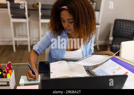 Busy biracial woman working at home sitting at table with laptop reading document and making notes Stock Photo