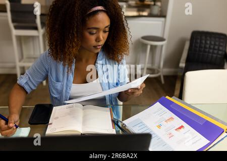 Busy biracial woman working at home sitting at table with laptop reading document and making notes Stock Photo