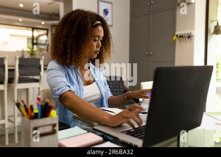 Busy biracial woman working at home sitting at table using laptop Stock Photo