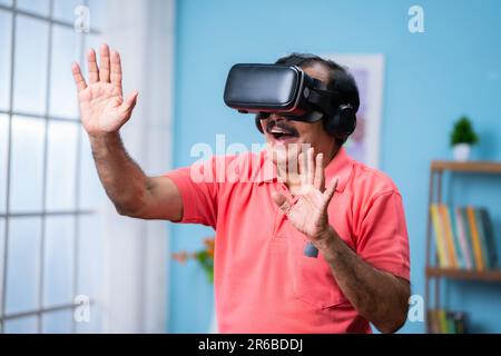 Happy indian senior man with VR or virtual reality headset experiencing metaverse at home - concept of imagination, futuristic and technology Stock Photo