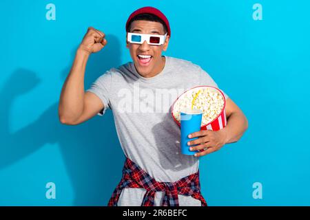 Portrait of crazy funky handsome guy raise fist hold soda drink popcorn watch movie 3d glasses isolated on blue color background Stock Photo