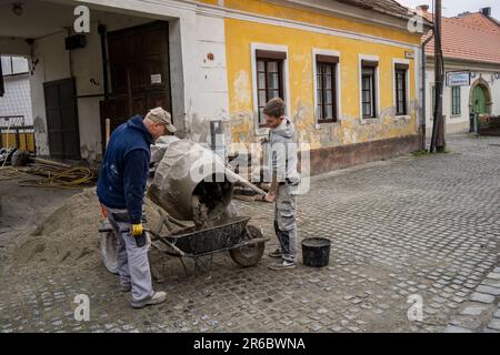 Szentendre, Hungary - November 30th, 2022: Two construction workers pouring cement into a wheelbarrow on a village street. Stock Photo