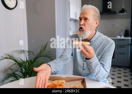 Angry annoyed bearded mature elderly male tasting slice of pizza with suspicion and with grimace of displeasure putting aside bad quality food sitting Stock Photo