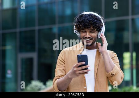 A young male Hispanic student is standing on the street near the campus wearing headphones with a phone in his hands. Listens to music, a podcast, talks on a video call. Stock Photo