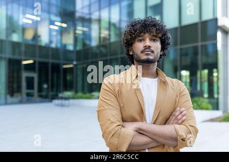 Portrait of a confident and successful young professional programmer, hispanic man standing outside an office center, crossing his arms over his chest and looking at the camera. Stock Photo