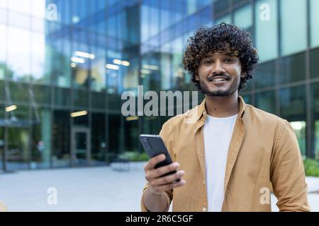 Close-up photo. Portrait of a young hispanic male computer programmer standing outside an office center and using the phone. Smiling looking at the camera. Stock Photo